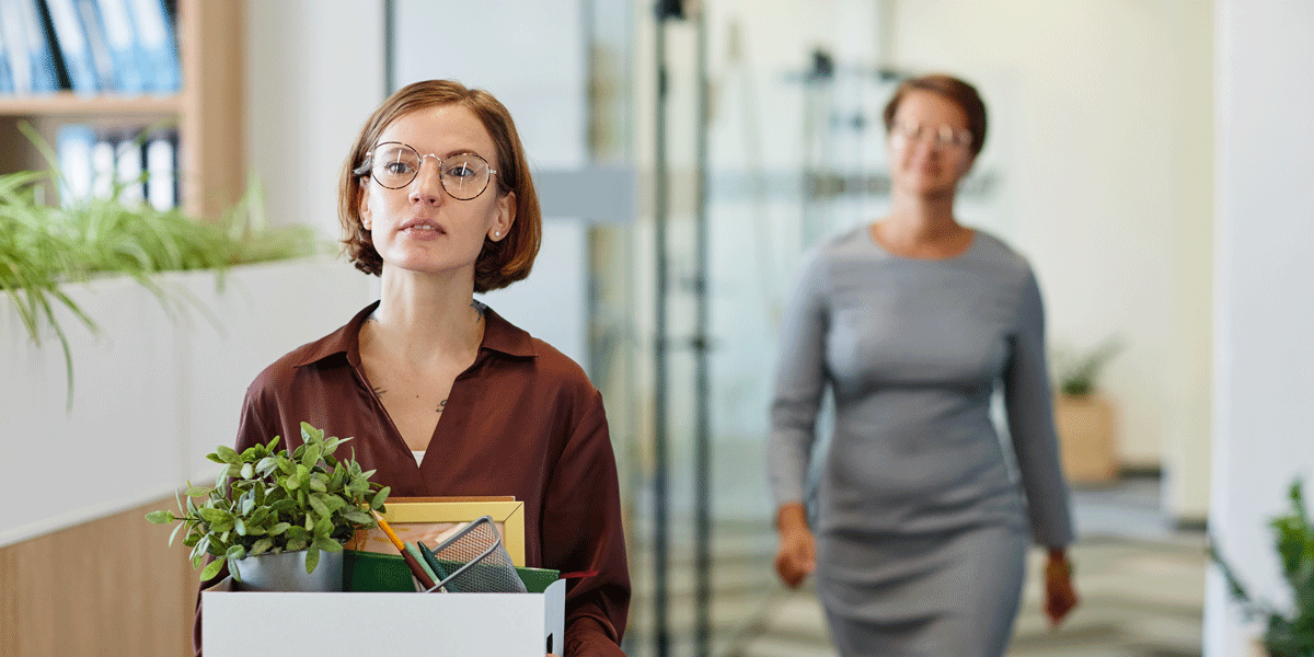 A young woman carrying a box of things from her desk. An older woman walks behind her.